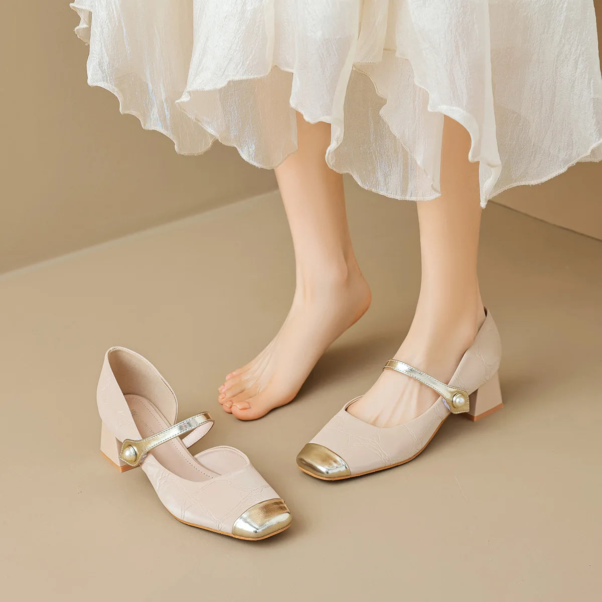Beige high-heeled shoes with gold accents worn by a person in a white skirt on a beige background