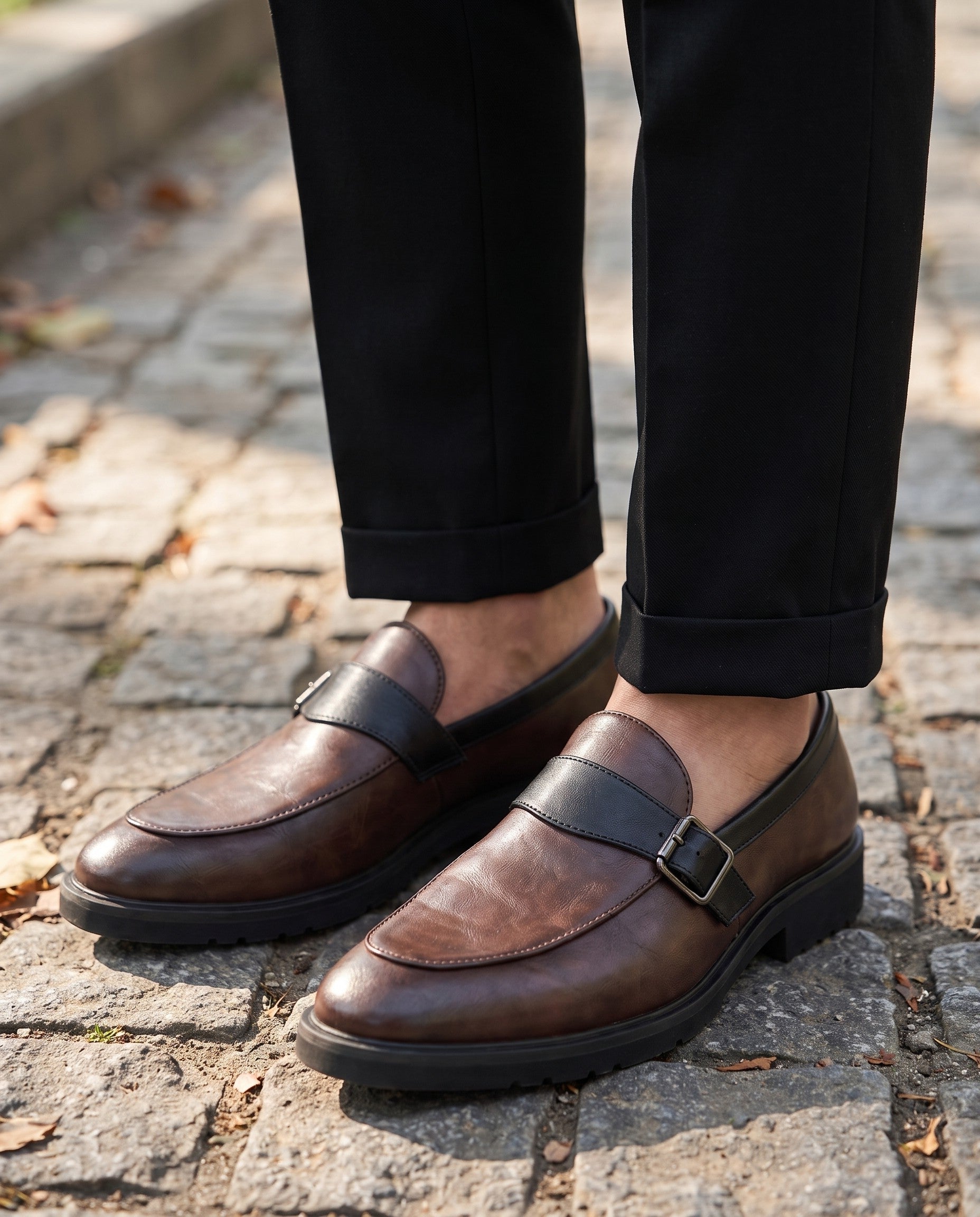 A close-up of a person wearing dark brown leather penny loafers with black straps and buckles, paired with black trousers, standing on a cobblestone path outdoors.